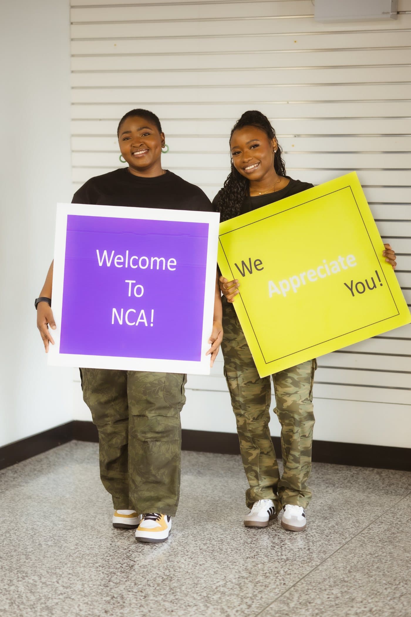 Two NCA volunteers smiling and holding signs that read Welcome to NCA and We appreciate you